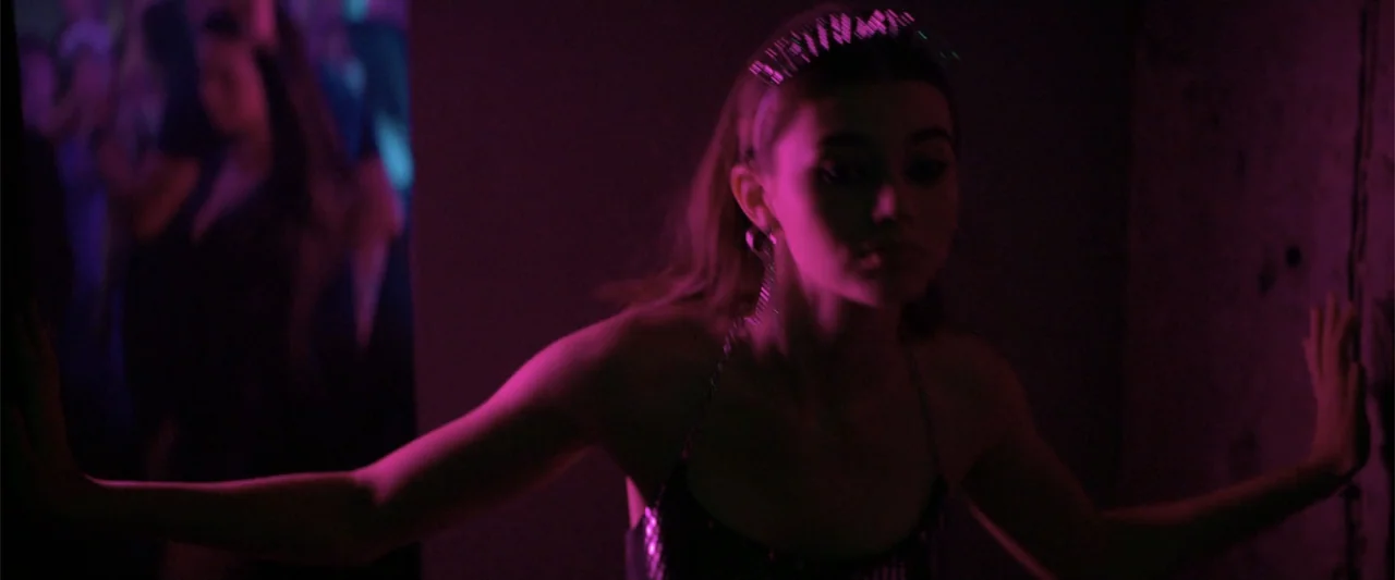 Marked Men: Rule + Shaw (2025) movie still: medium — A dark, enclosed space is bathed in intense purple and pink light. A young woman with a…