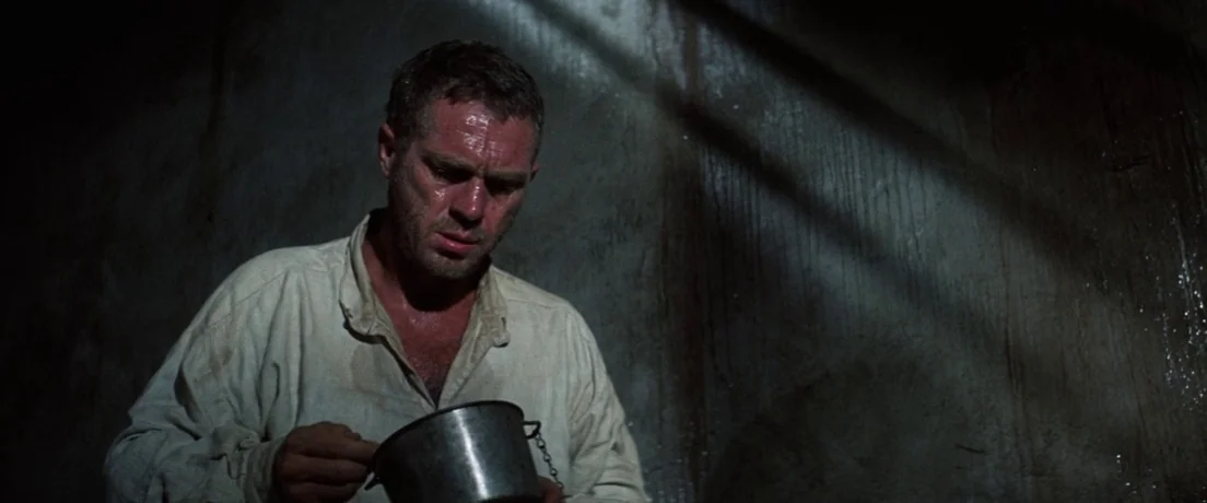 Papillon (1973) movie still: medium — A dark stone cell with damp walls and bars casting sharp diagonal shadows. A man in a disheveled shirt holds a…