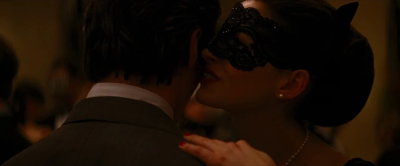 The Dark Knight Rises (2012) movie still: closeup — A dimly lit, formal event space with guests in evening wear and masquerade masks. A woman leans…