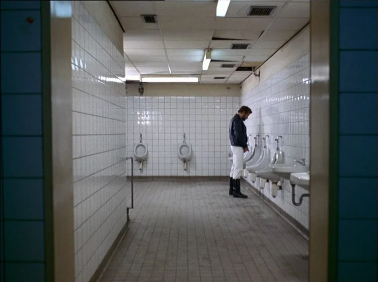 Satan’s Brew (1976) movie still: wide — A utilitarian public restroom features clinical white square wall tiles and drab floor tiles. A man stands…