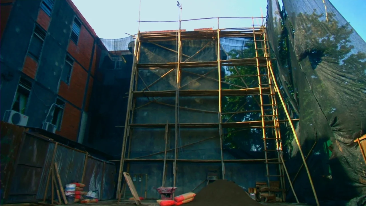 Merantau (2009) movie still: wide — A construction site features a tall bamboo scaffold erected against a concrete building exterior. A pile of sand…