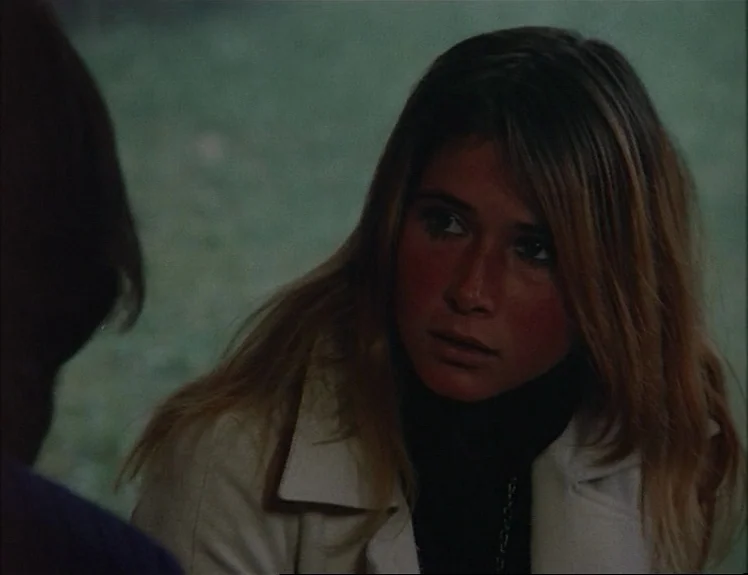 Claire’s Knee (1970) movie still: closeup — A woman faces a companion in a grassy field during the daytime. The woman wears a white coat and looks…