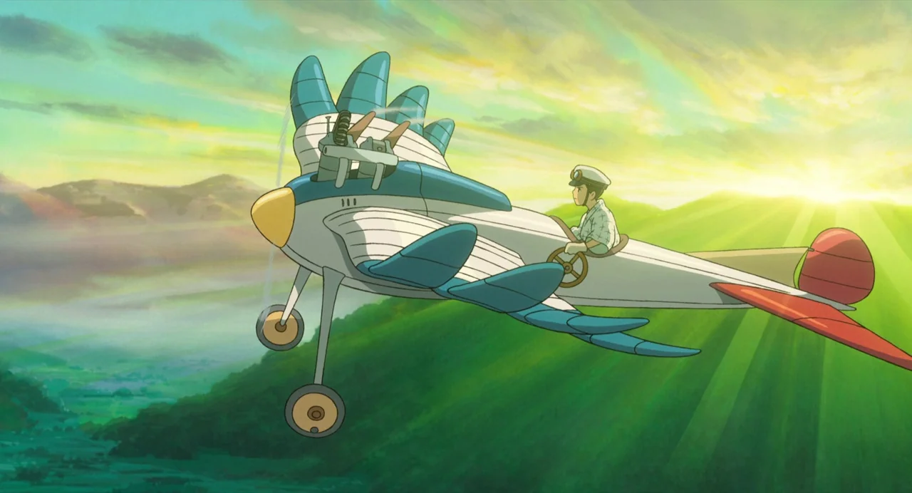 The Wind Rises (2013) movie still: wide — A fantastical airplane with blue wings flies above a vast landscape of green mountains. A young male pilot…