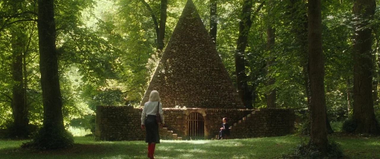 Knife+Heart (2018) movie still: wide — A stone pyramid structure sits in a dense clearing surrounded by mature deciduous trees. A blonde woman walks…