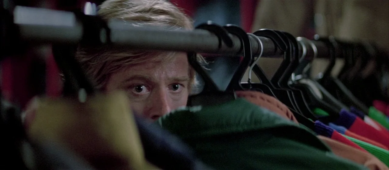 Three Days of the Condor (1975) movie still: closeup — A close view through a dense rack of assorted winter coats on black hangers. A man with blond…