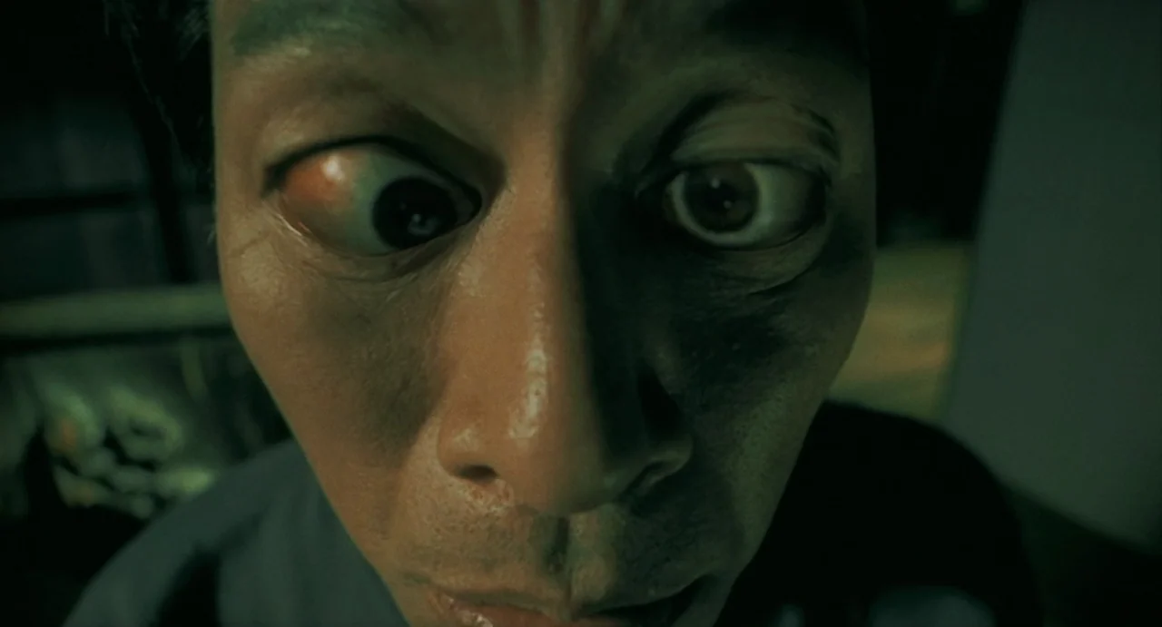Uzumaki (2000) movie still: closeup — A dark, dimly lit indoor space with indistinct textures visible in the background. An East Asian man faces the…