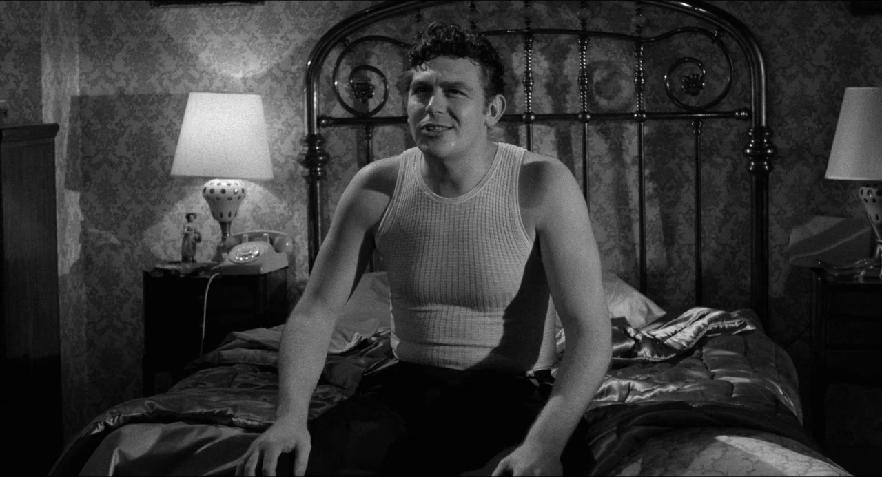 A Face In The Crowd (1957) movie still: medium — A furnished space with floral wallpaper, a metal bed frame, and two bedside tables. A young man…