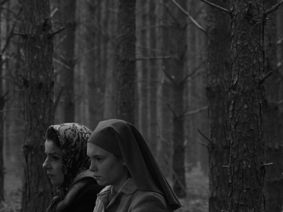 Ida (2013) movie still: medium — A dense, uniform stand of tall pine trees forms the backdrop of the frame. Two women stand in the foreground,…