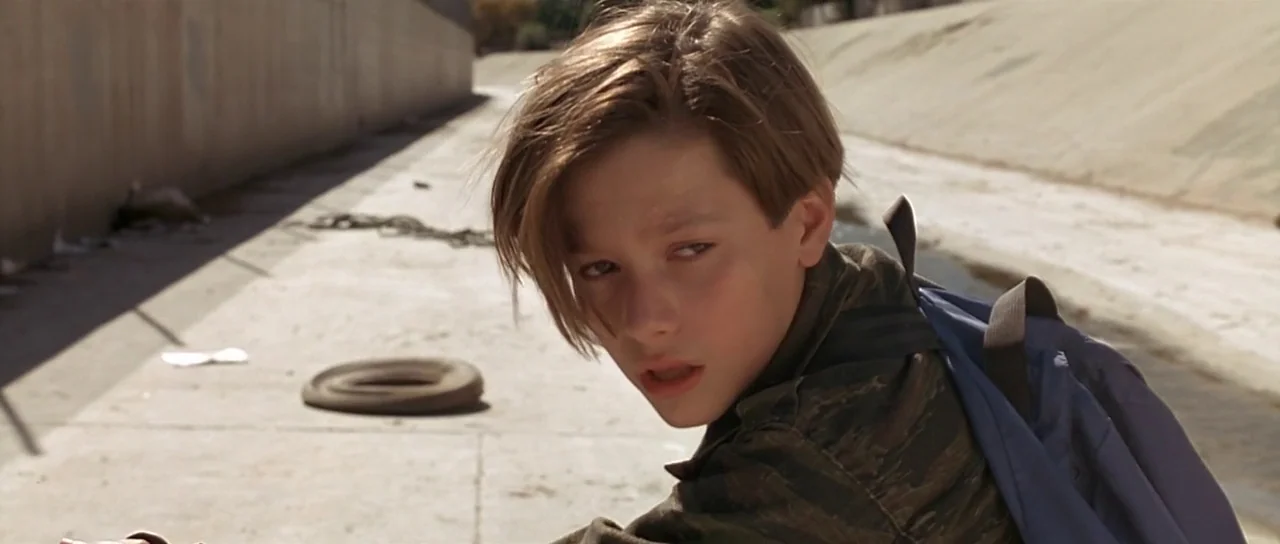 Terminator 2: Judgement Day (1991) movie still: medium — A wide, dry concrete canal with slanted walls under bright daytime sun. A teenage boy in a…
