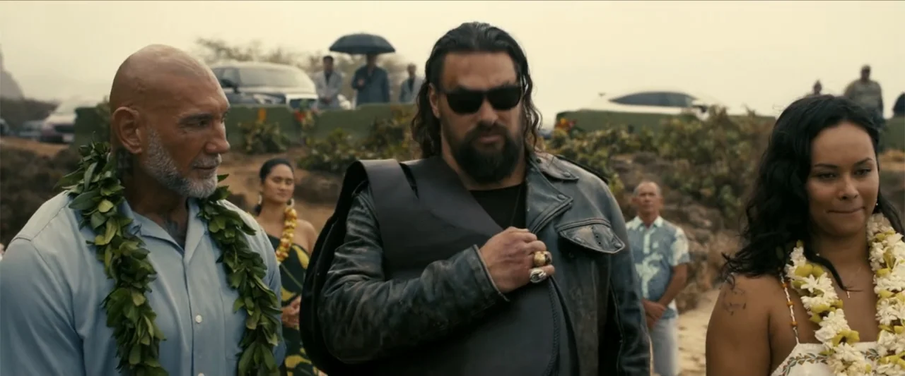 The Wrecking Crew (2026) movie still: medium — An outdoor gathering takes place near a rocky shore with vehicles parked in distance. Three primary…