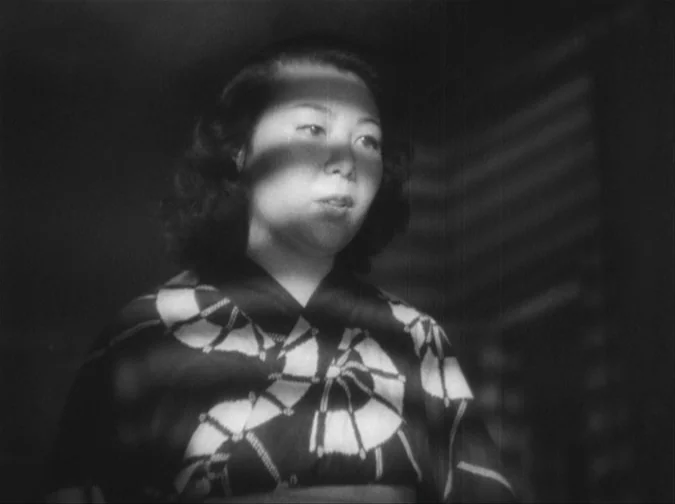 Drunken Angel (1948) movie still: medium — A dark room is illuminated by light filtering through horizontal window blinds. A woman in a patterned…