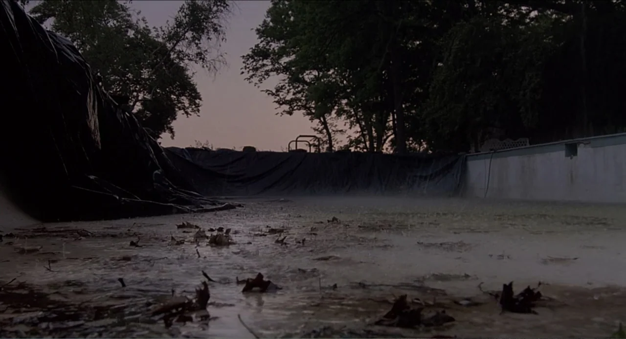 Rain Man (1988) movie still: wide — A neglected outdoor swimming pool is covered in dark plastic sheeting at twilight. Rain falls onto the…