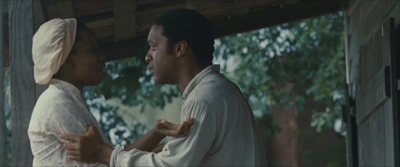 12 Years a Slave (2013) movie still: medium — A wooden porch overlooks a blurred green outdoor space in a rural 19th-century setting. A man in a…