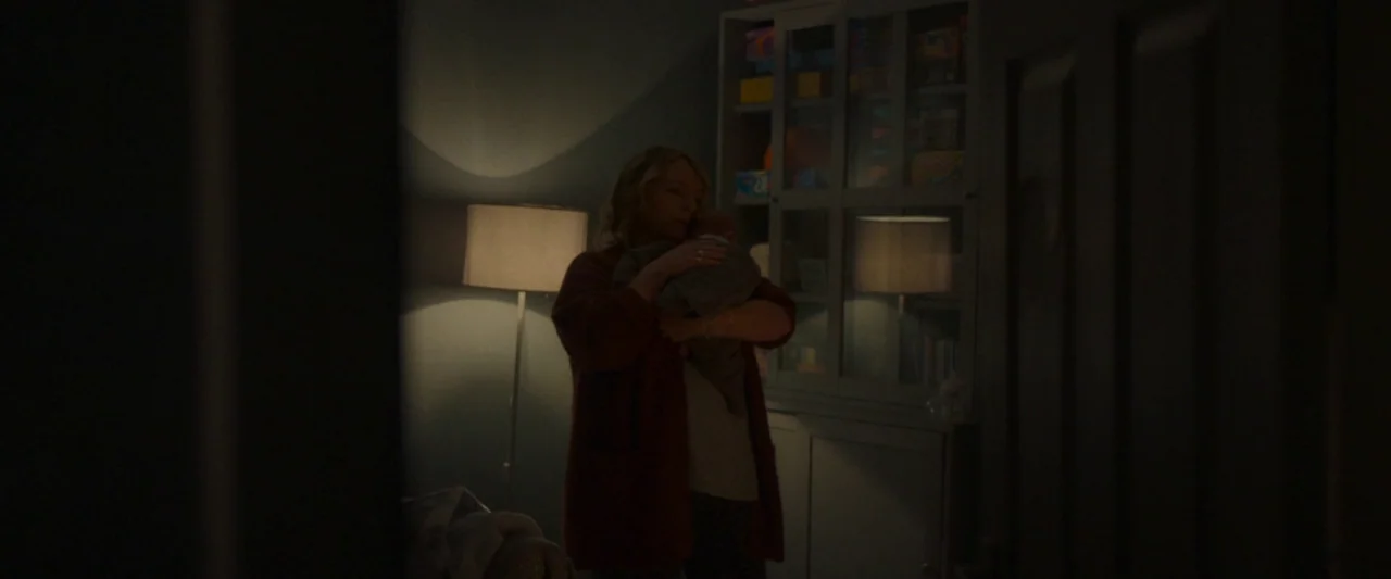 Ali & Ava (2021) movie still: medium — A dimly lit room contains a floor lamp and a tall glass-fronted cabinet. A woman stands in the low light…