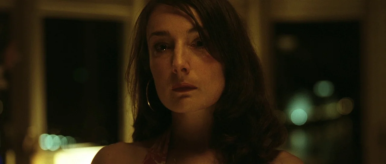 Bullhead (2011) movie still: closeup — A dark, dimly lit room with blurred lights visible in the background. A woman faces the camera directly in a…