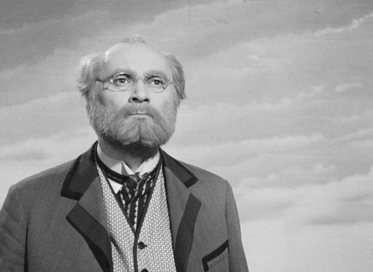 Invention For Destruction (1958) movie still: medium — An older man stands before a painted canvas background depicting a cloudy sky. He gazes…