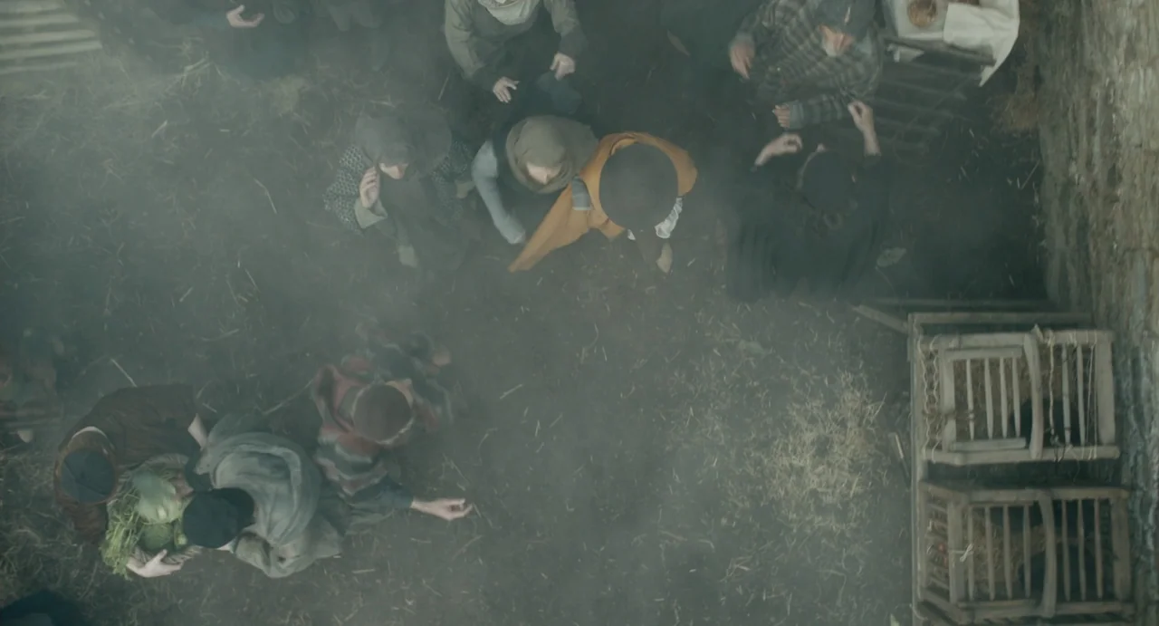 Catherine Called Birdy (2022) movie still: wide — A muddy, fog-filled open courtyard is populated by figures in rustic medieval clothing. People…