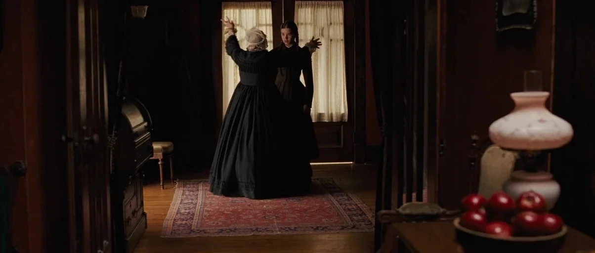 True Grit (2010) movie still: wide — A dimly lit 19th-century parlor features dark wood furniture and a red patterned rug. An elderly woman standing…