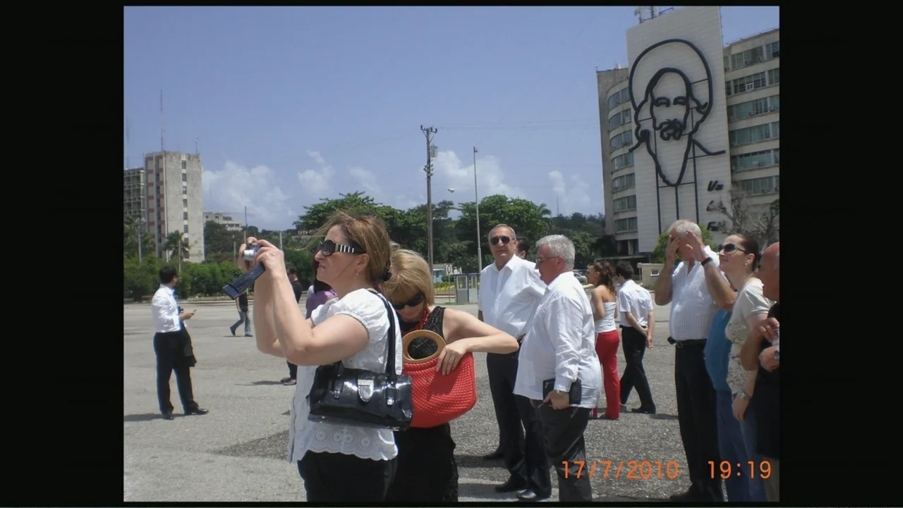 Letters to Max (2014) movie still: medium — A sunny public plaza features a large building with a mural of Camilo Cienfuegos. A small group of…