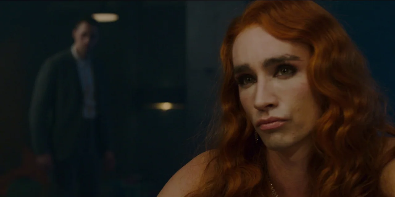 Mute (2018) movie still: closeup — A dimly lit interior space contains a person in the foreground and another distant. The foreground subject with…
