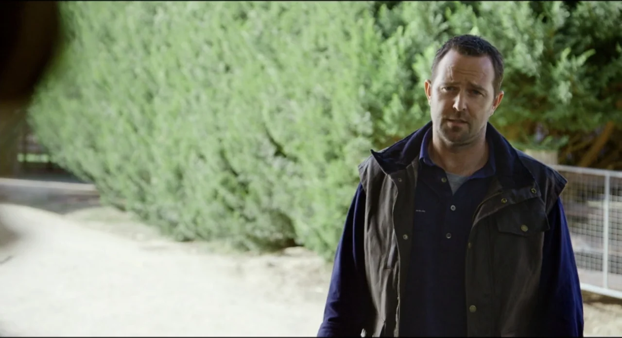 Ride Like a Girl (2019) movie still: medium — A man stands outdoors in front of a dense green hedge and wire fencing. He is wearing a dark polo and…