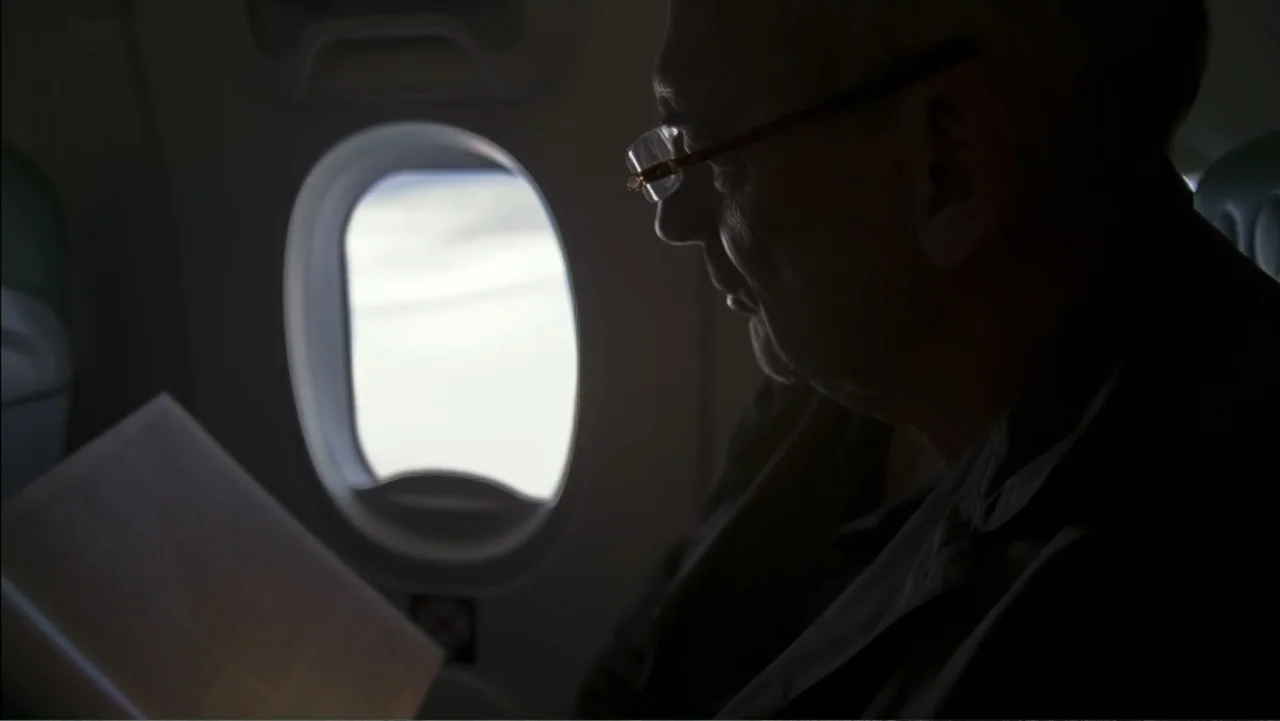 REM (2019) movie still: closeup — An interior of an airplane cabin shows a passenger sitting by the window. The subject reads a document in the low…