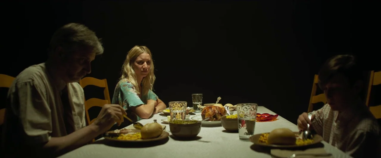The Mad Hatter (2021) movie still: medium — A family sits around a dining table against a pitch black background. The subjects are eating dinner…