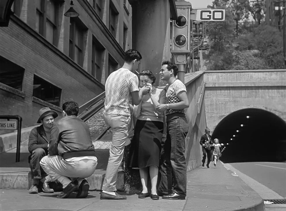 M (1951) (1951) movie still: fullbody — A group of young adults congregates by a concrete tunnel entrance next to buildings. The individuals stand…