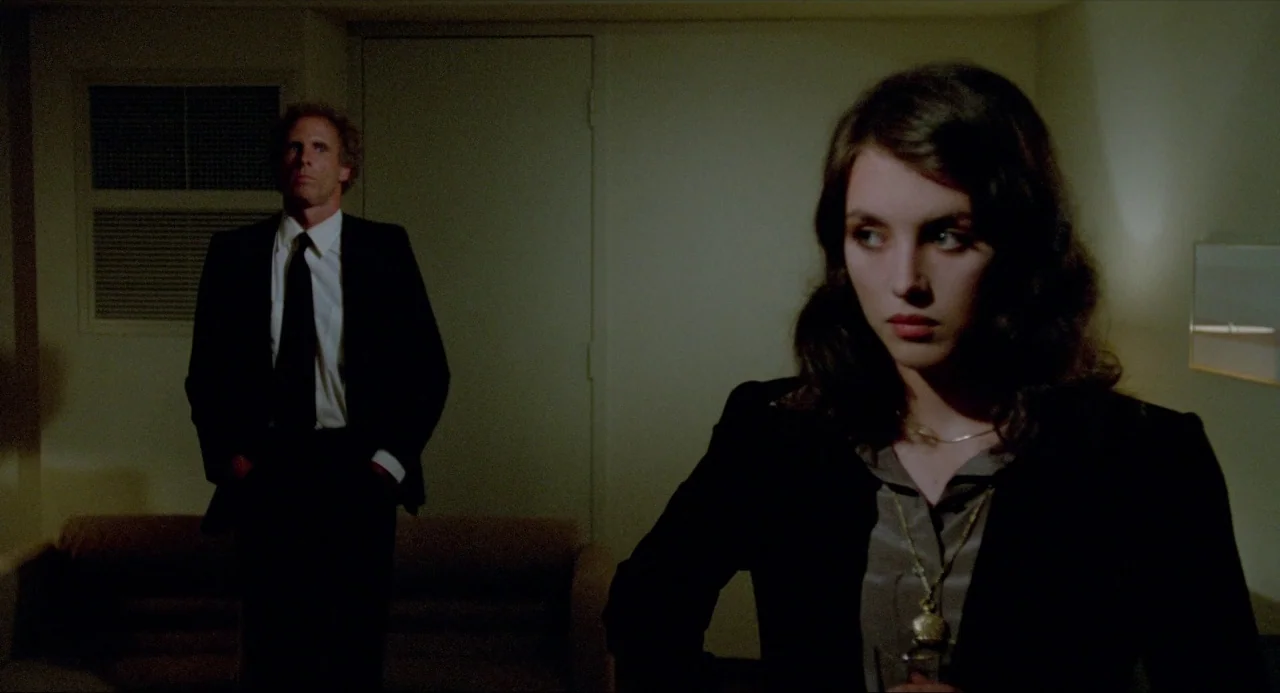 The Driver (1978) movie still: medium — A dimly lit room with plain walls and an air conditioning unit. A woman stands in the foreground while a man…