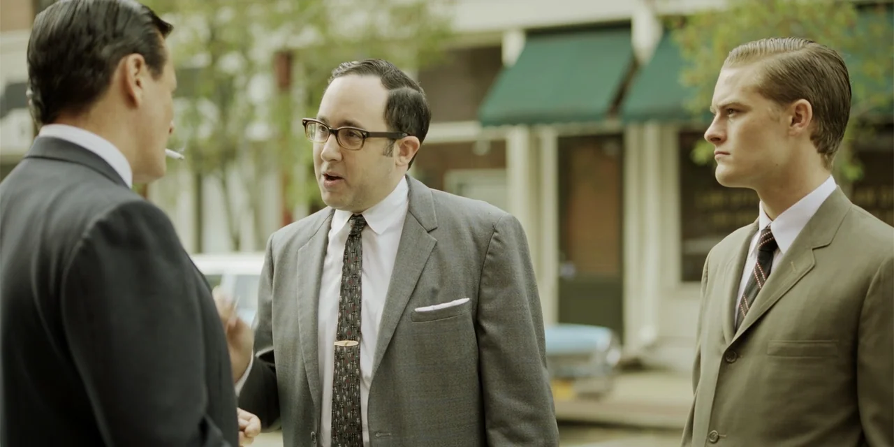 Green Book (2018) movie still: medium — A group of men in 1960s business attire gather on a concrete sidewalk. They are engaged in a mid-day…