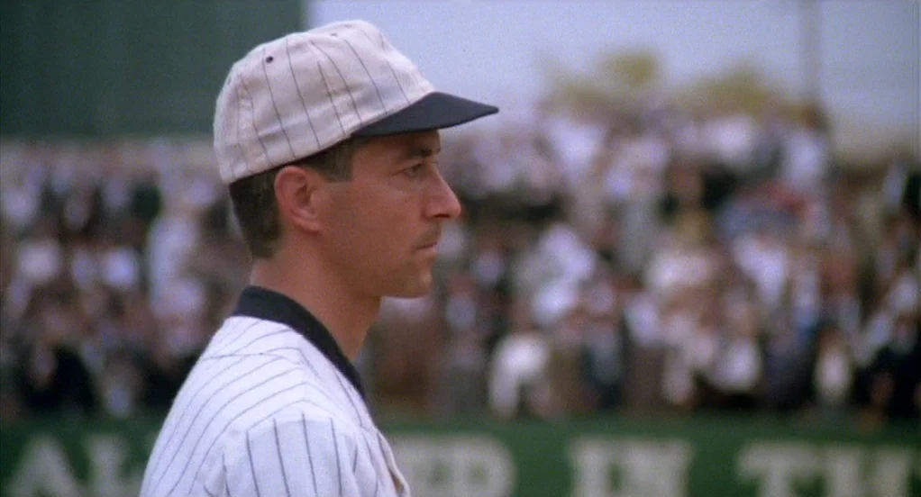 Eight Men Out (1988) movie still: closeup — A baseball player in vintage pinstriped gear stands on a field during a game. A blurred crowd of…