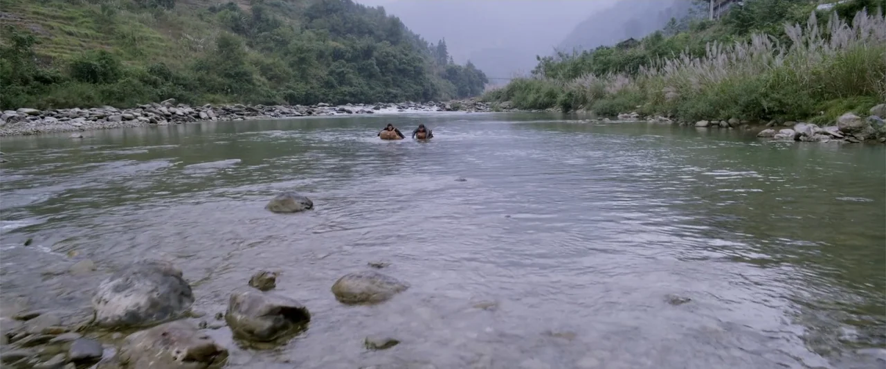 Skiptrace (2016) movie still: wide — A wide, shallow mountain stream flows over rocks with forested hills in the background. Two men wade slowly…