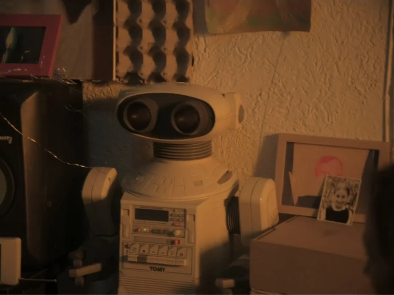 History and Geography (2024) movie still: medium — A cluttered interior shelf holds a vintage plastic robot toy next to framed photographs. The…
