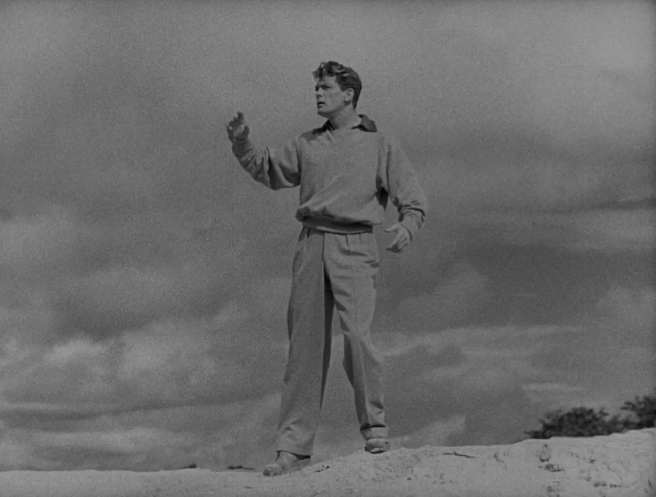 Orpheus (1950) movie still: fullbody — A man stands atop a sandy dune beneath an expansive, cloud-filled sky. He gestures with his right arm…