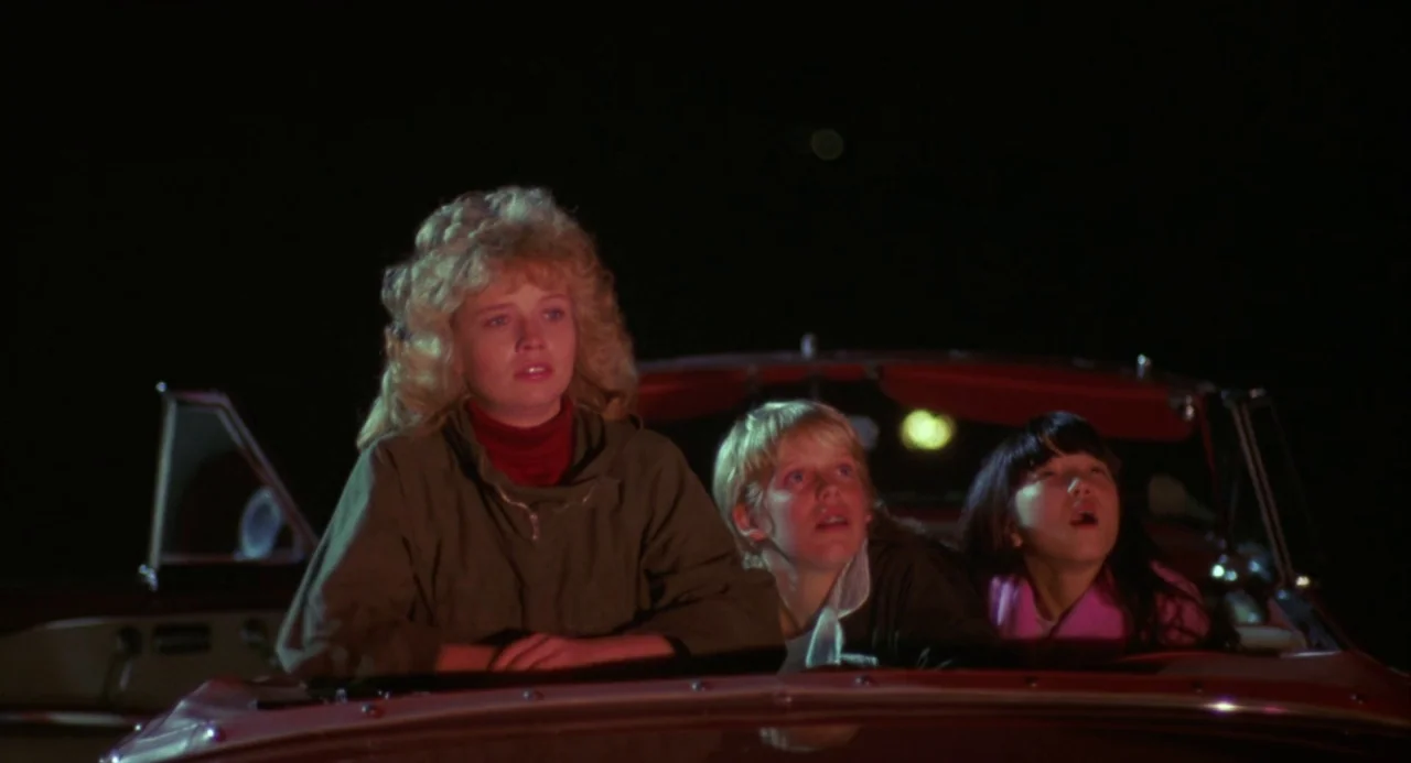 Night of the Comet movie still: medium — A night scene shows three people seated together inside the front of a vehicle. They look upward at…