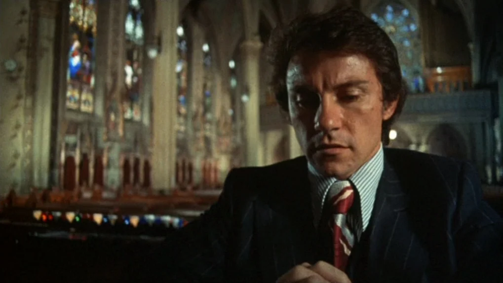 Mean Streets (1973) movie still: medium — A man stands in the foreground of a large, empty stone church nave. Stained glass windows and rows of…