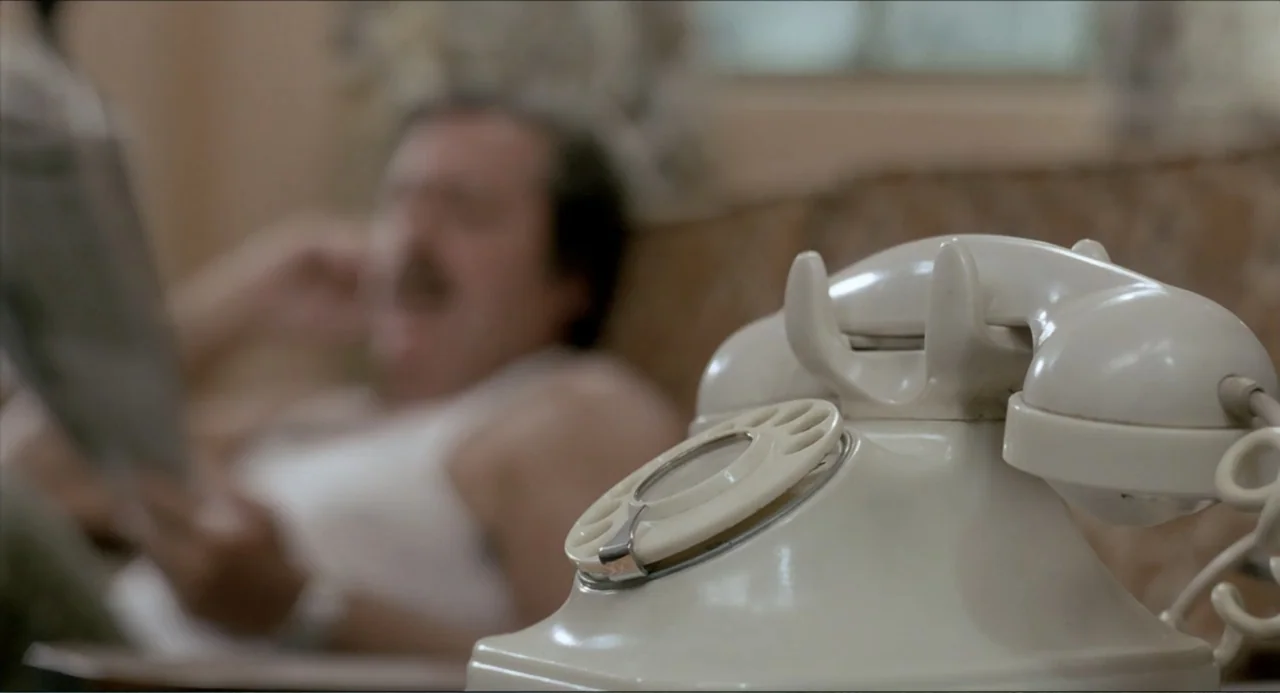 Waiting for the Hearse (1985) movie still: closeup — A domestic interior features a beige patterned sofa and soft daylight. A man is partially…