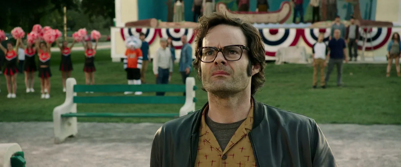 It Chapter Two (2019) movie still: medium — A public park features a green wooden bench and a small performance stage behind. A man with glasses…
