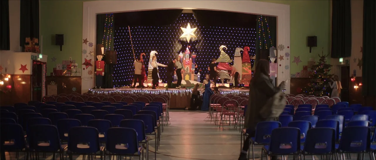Anna and the Apocalypse (2018) movie still: wide — A community hall interior features rows of blue plastic chairs facing a decorated stage. Several…