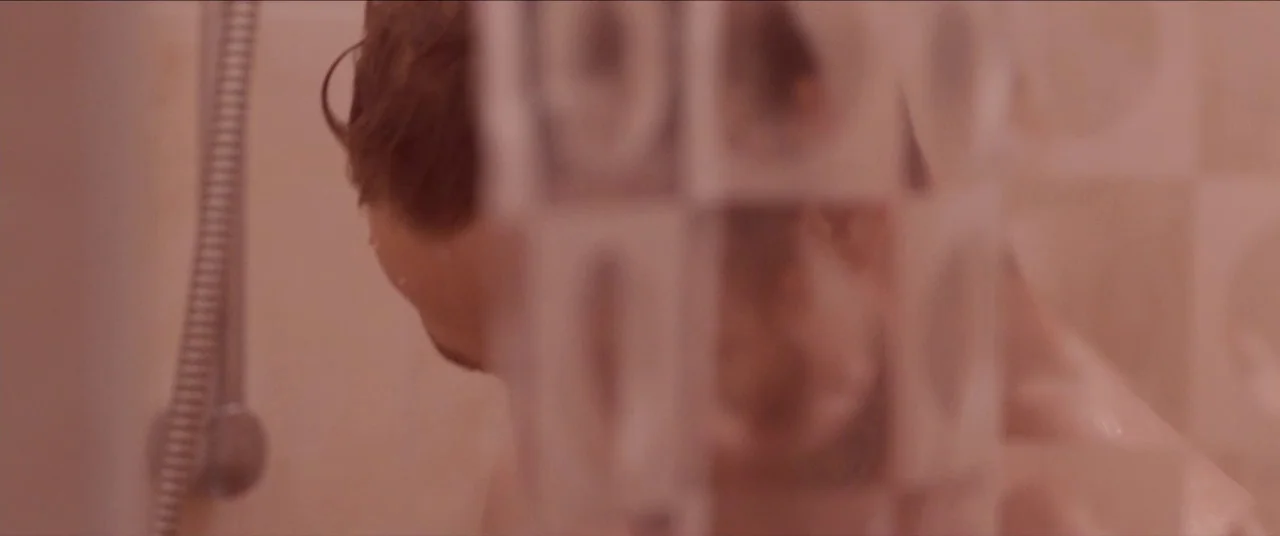 Simon Killer movie still: closeup — A man stands in a shower viewed through a textured, translucent plastic curtain. Water droplets are visible on…