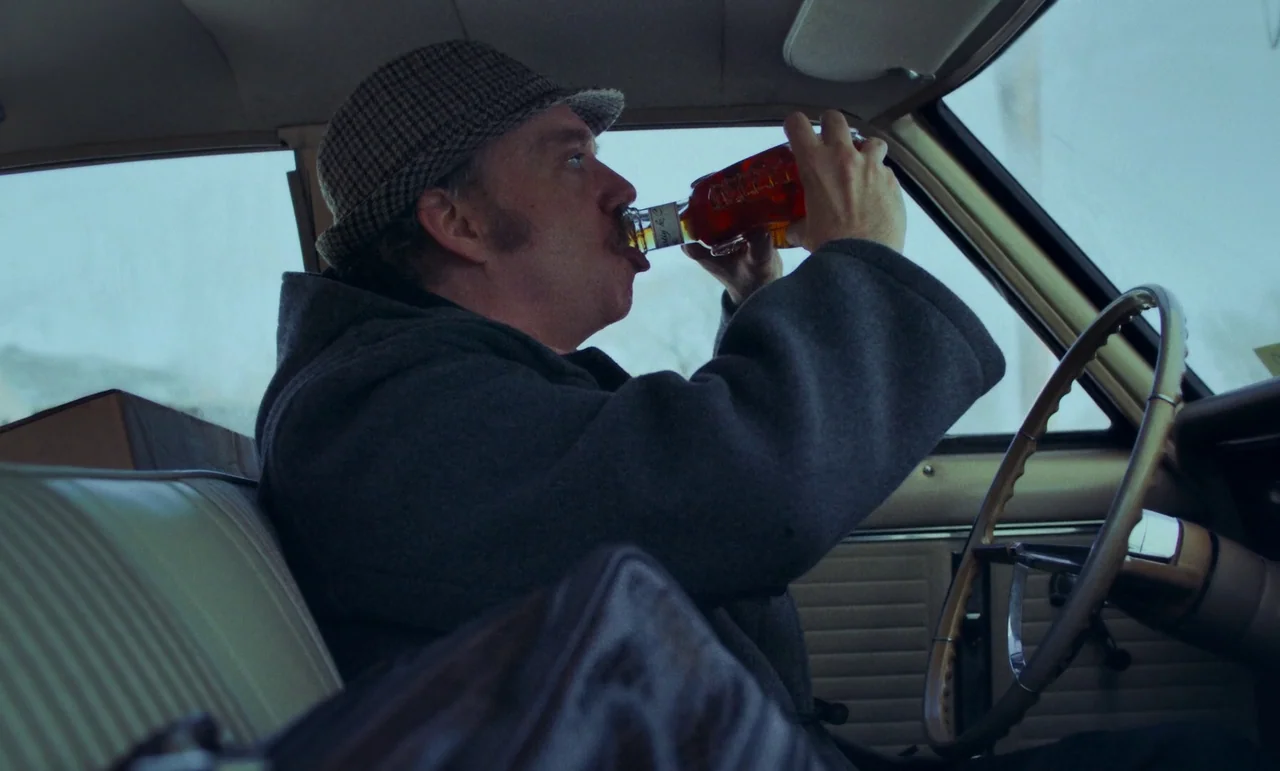 The Holdovers (2023) movie still: medium — The interior of a vintage car features a beige bench seat and steering wheel. A man wearing a flat cap…