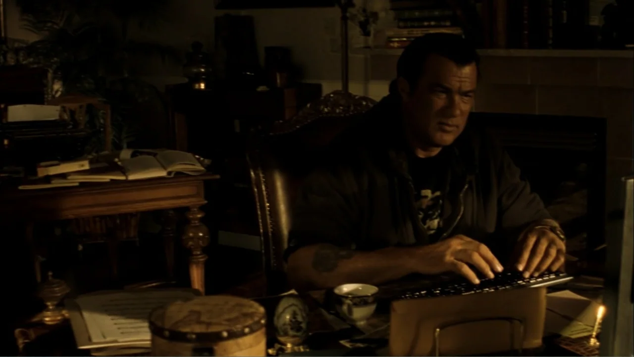 Driven to Kill (2009) movie still: medium — A dark home office contains a wooden desk with a computer and assorted books. A man sits at the desk and…