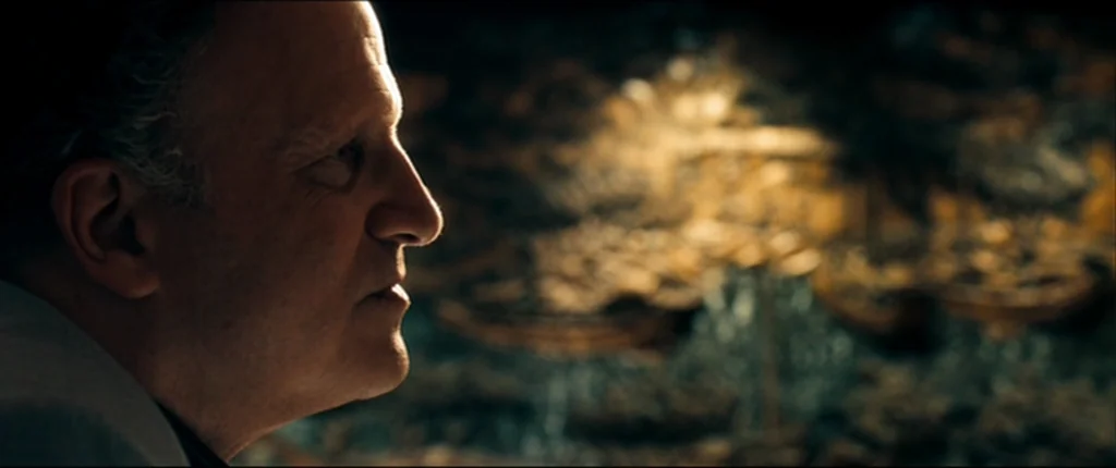 Drive (2011) movie still: closeup — An older man looks at a blurred collection of golden mechanical gears in shadow. Sharp lighting highlights the…