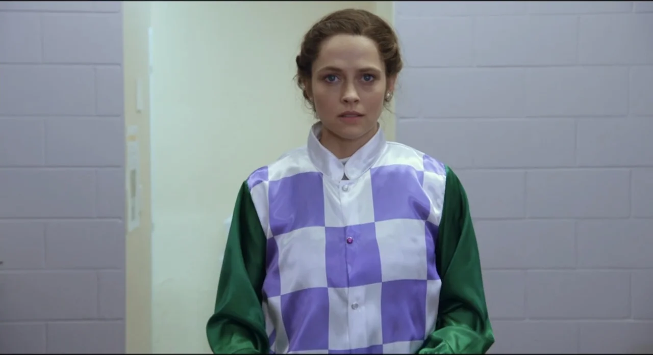 Ride Like a Girl (2019) movie still: medium — A woman stands in a functional room with white cinder block walls. The subject wearing a jockey…