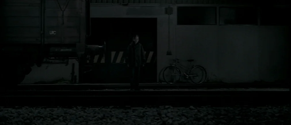 Time Of The Wolf (2003) movie still: wide — A dark night scene shows a child standing on gravel beside railway tracks. A large freight train car…