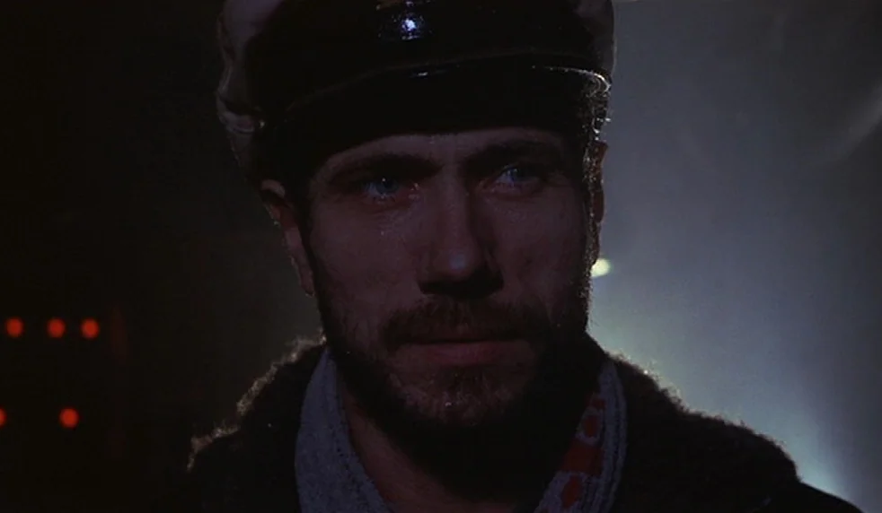 Das Boot (1981) movie still: closeup — A man in a captain's hat stands in a dark, poorly lit space. He gazes off-camera with a beard and intense eyes.