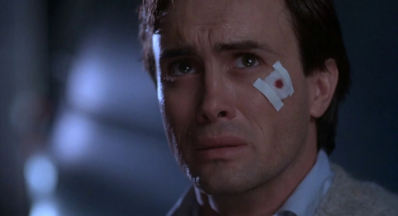 From Beyond (1986) movie still: closeup — A dimly lit room with a dark, indistinct background. A man with a blood-spotted bandage on his cheek gazes…