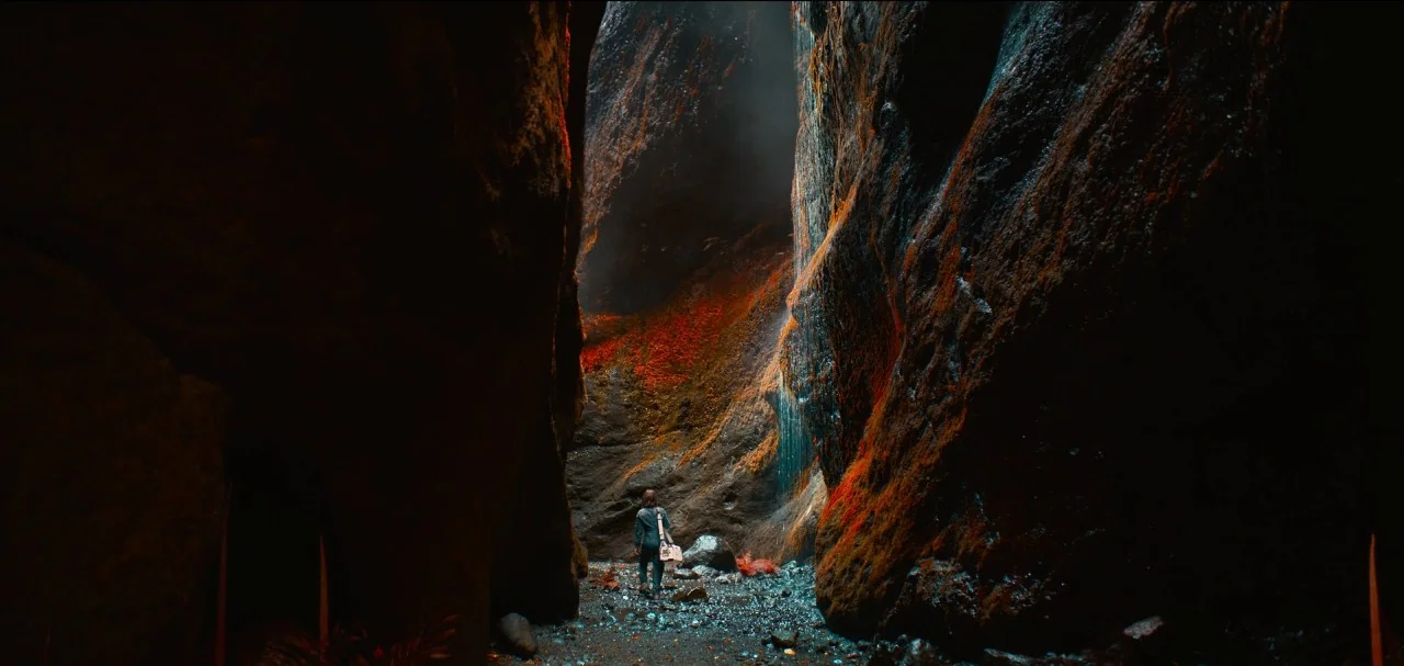 The Midnight Sky (2020) movie still: wide — A narrow, dark canyon has towering rock walls covered in patches of bright orange moss. A thin waterfall…