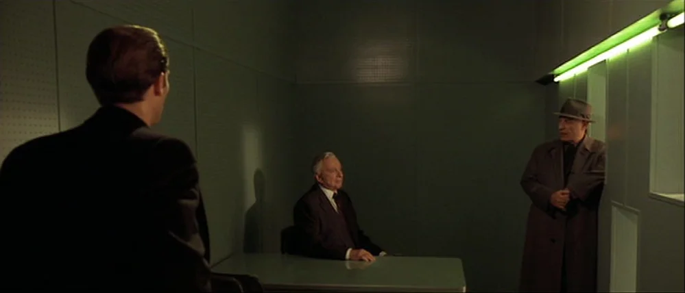 Gattaca (1997) movie still: wide — An institutional room features flat grey walls with a thin green light strip. Three men stand and sit inside the…