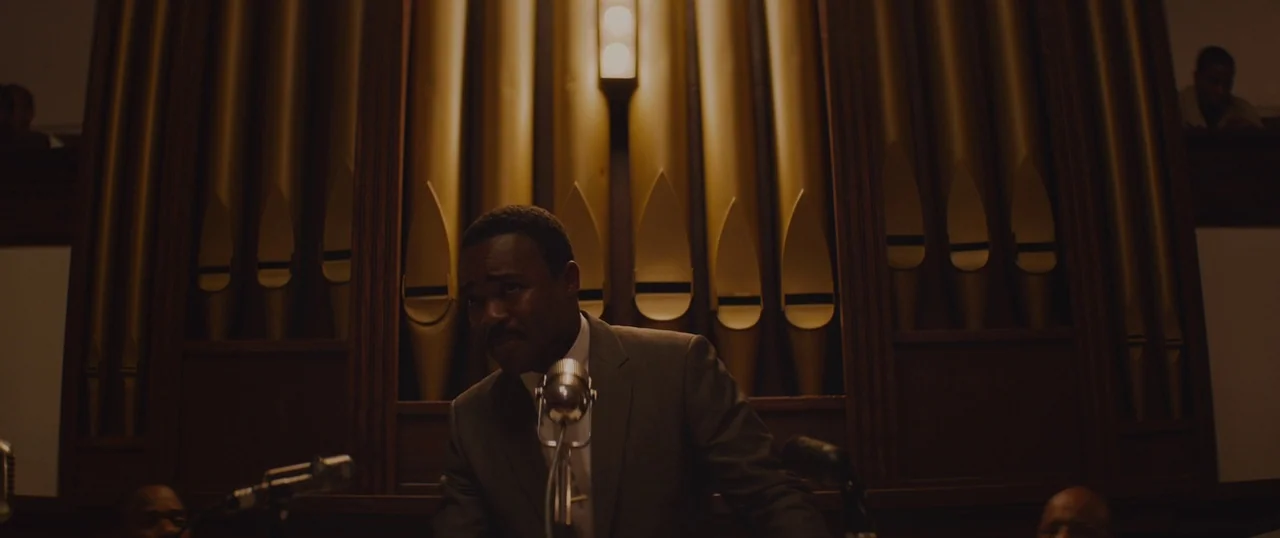 Selma (2014) movie still: medium — A man stands behind a vintage microphone in front of a wooden pipe organ. The organ pipes are gold-colored and…